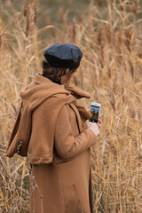 Portrait of young  woman in a black shiny beret and a beige wool coat photographs for his blog,   posing against  the autumn field, countryside. Blogger work and content creation concept