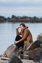 A romantic walk of a curly-haired woman in  beige wool coat   posing on stone in sea. The concept of female freedom, emancipation and love