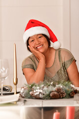 Happy looking mature asian woman with a Santa hat sitting at a table with Christmas decorations. Christmas holiday and celebration concept.