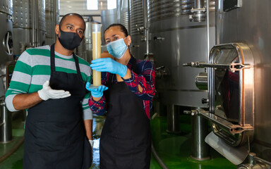 Man and woman workers of winery in protective masks checking wine production process at fermentation tank