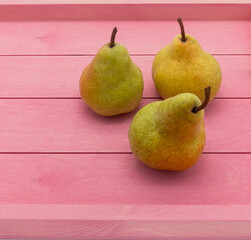 Three ripe yellow pears are on a pink wooden tray. There are specks on the peel. Angle view.