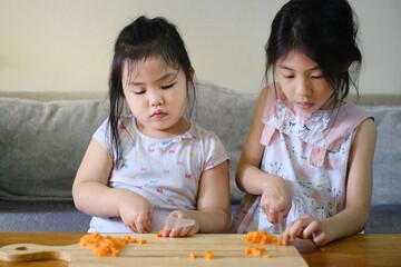 Two cute young Asian girls are helping their mother prepare their dinner by chopping raw carrots on a table using plastic knifes.