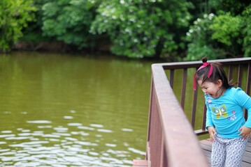 A cute young Asian girl, standing on a bridge by a pond, looking down at the water and feeling excited after seeing a turtle. Jumping up and down with joy.