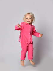 Frolic barefooted curly blonde baby girl in pink warm comfortable jumpsuit jumps looking at camera swinging waving arms