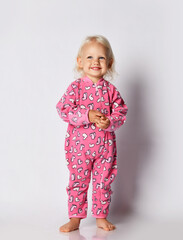 Smiling barefooted blonde baby kid girl in pink warm comfortable jumpsuit stands over grey wall background and look up