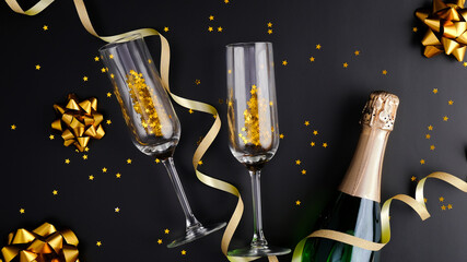 New Year Eve celebration concept. Black Christmas background with champagne bottle, drink glasses, golden confetti stars. Flat lay, top view.