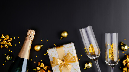 Christmas champagne bottle, gift box, glasses, golden confetti, balls decorations on dark background. Flat lay, top view. Xmas party, New Year celebration concept.