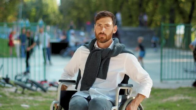 At sunlight portrait handsome disabled man in wheelchairat look at camera serious in park. Paralysis disability positive handicapped. Slow motion