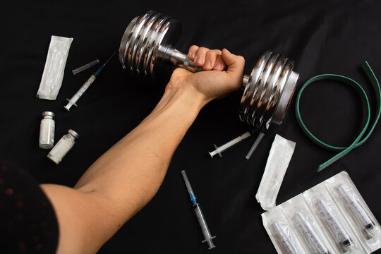 man holds dumbbell with tense hand on black background, on which there are used syringes and packages, tourniquet, ampoules. The concept of sports and drugs, pharmacology. Soprt against drug