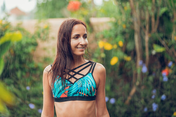 Portrait of a beautiful tanned, slender woman in a swimsuit, posing under a refreshing tropical rain. Plants and flowers on the background. Summer vacation and travel concept