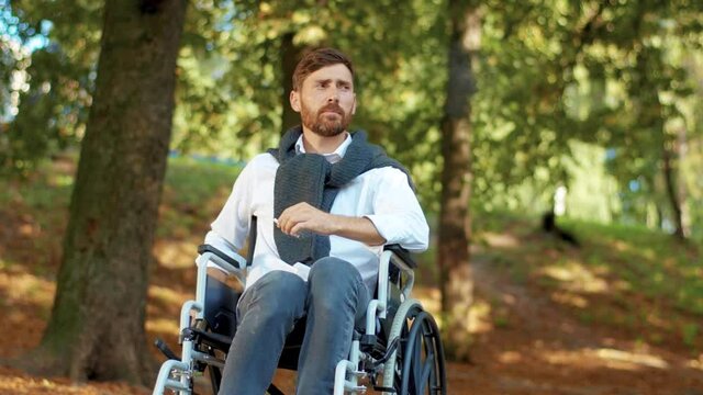 Serious young man in wheelchair smoking cigarette in beautiful park at sunlight. Disability walking bearded handsome. Portrait. Slow motion