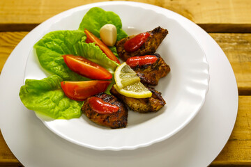 Pieces of grilled meat with tomato, salad and lemon are laid out on a plate.