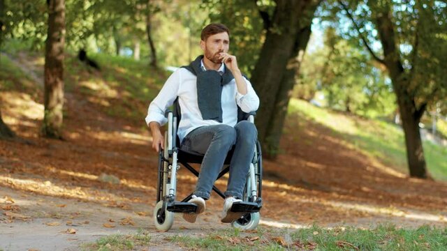 Sad attractive young man in wheelchair smoking cigarette in beautiful park at sunlight. Disability walking bearded handsome. Portrait. Slow motion