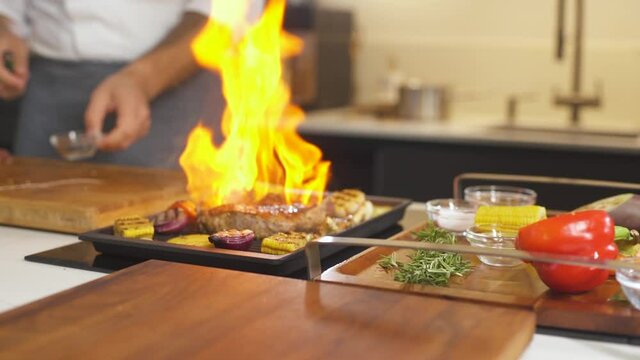 Cooking Meat Steak With Vegetables At Modern Kitchen On Grill. Male Chef Preparing Flambe Grilled Dish With Fire At Restaurant. Flame In Frying Pan. Making Delicious Food. Processing Meal On Cuisine.