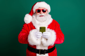 Photo of retired stylish grandpa grey beard hold telephone share post social network wear red santa x-mas costume coat gloves sunglass headwear isolated green color background