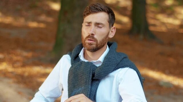 Close up face sad attractive young man in wheelchair smoking cigarette in beautiful park at sunlight. Disability walking bearded handsome. Portrait. Slow motion