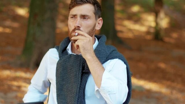 Face serious young man in wheelchair smoking cigarette in beautiful park at sunlight. Disability walking bearded handsome. Portrait. Slow motion