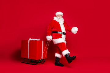 Photo portrait of man in santa claus costume pulling big wrapped gift box on sleigh isolated on vivid red colored background