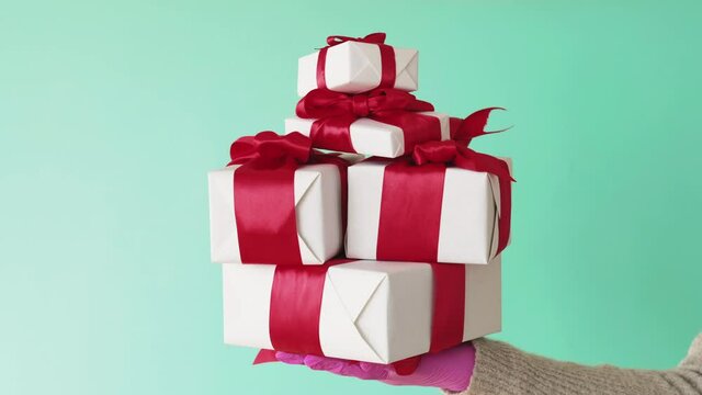 Pandemic Christmas gifts. COVID-19 New Year 2021. Quarantine winter holidays sale. Hands in pink protective gloves showing stack of white boxes with red ribbon bows isolated on blue background.