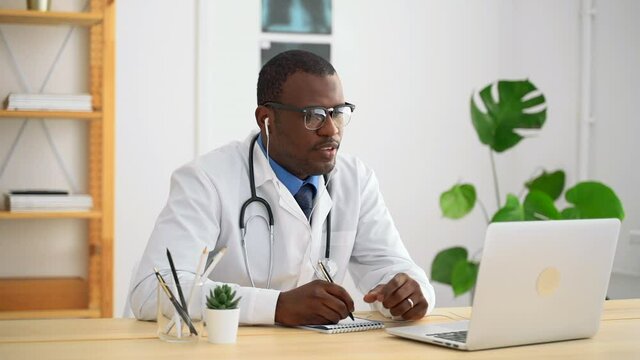 Young male doctor is writing during online consultation at table in modern clinic spbas. African American man taking notes and talking while looking at laptop screen at desk in bright room