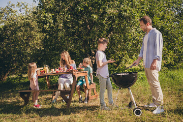 Family full outdoors gathering. People dad daddy cook roast grill bbq sausages boy talk conversation mom mommy sit table have meal under green garden tree