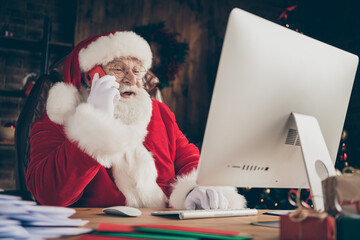 Photo white grey beard hair jolly holly santa claus sit table work computer check email look wish list letter call elf smartphone wear gloves cap headwear in x-mas christmas house indoors