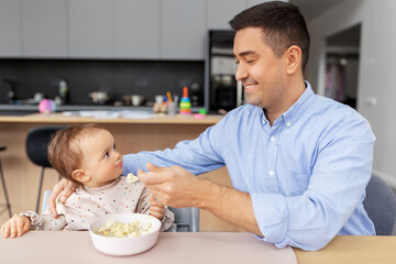 Obraz na płótnie Canvas family, food, eating and people concept - happy smiling middle-aged father feeding little baby daughter sitting in highchair with puree by spoon at home