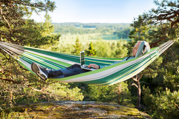 Obraz na płótnie Canvas A man working on a laptop while lying in a hammock in the woods. Self-isolation, freelancing, remote work and distancing. Top view of Scandinavian landscape 