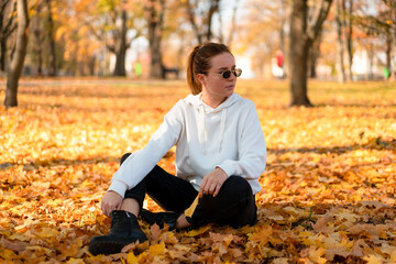 Woman in a white sweater with a hood sits on ground in the park and holding a leaf in her hands