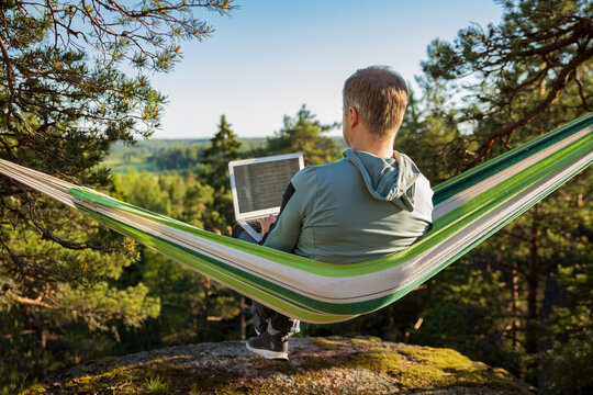 A Man Working On A Laptop While Lying In A Hammock In The Woods. Self-isolation, Freelancing, Remote Work And Distancing. Top View Of Scandinavian Landscape 