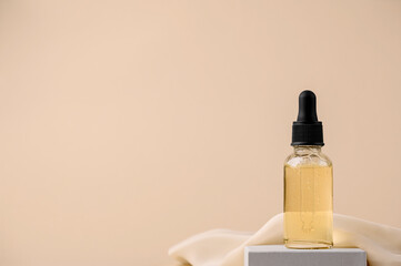 Serum in a glass bottle with a pipette on a gray stand with a silk cloth. Essential oil for the care of female skin. Skin care, moisturizing beauty product on a beige background. Copy space