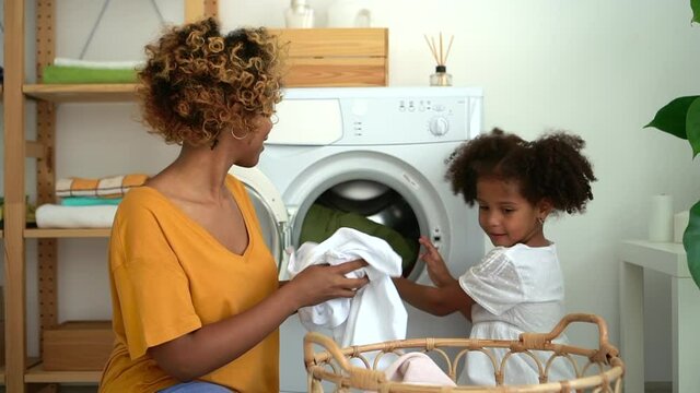 Young mother and little daughter putting clothes in washing machine in laundry room at home spbd. American African woman and cute girl talking with smiles and loading appliance while sitting on floor