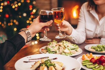 Close up of hands holding wine glasses and clinking against the backdrop of bright Christmas tree and burning fireplace, couple celebrating at Christmas table