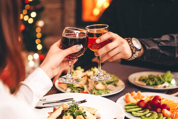 Close up of hands holding wine glasses and clinking against the backdrop of bright Christmas tree and burning fireplace, couple celebrating at Christmas table