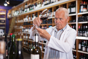 Confident elderly male winemaker holding glass of red wine, checking it in wine store..