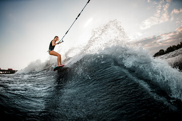 view of young woman on surf style wakeboard who holds on to rope and rides on wave from boat