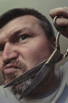 An adult man cuts his own beard with old huge scissors.