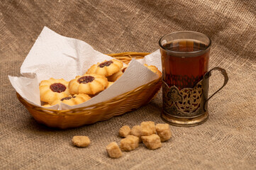 Homemade pastry cookies with jam and a faceted glass of tea in a vintage Cup holder on a background of homespun fabric with a rough texture, close-up, selective focus.