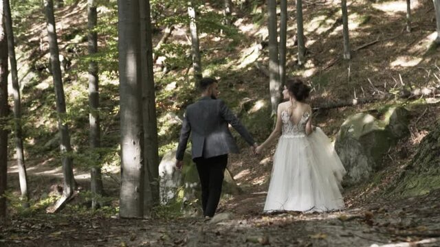Among the autumn forest with brown and green leaves on the trail, a guy in a jacket and black pants walks by the handle with a girl in a white lush long dress, a couple in love walks away