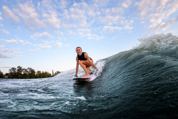 smiling young woman sit with bent knees on surf style wakeboard and rides on wave