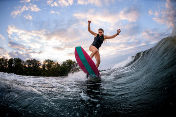 energetic young woman stands on surf style wakeboard and ride down on wave