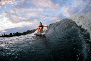 view on happy woman riding wave while sitting on surf style wakeboard with arms outstretched to the sides