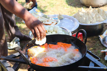 Cooking Of Various Types Of Deep Fried Indian Egg Bajji Or Pakodas In Hot Boiling Oil And Hands Of Man Dropping Uncooked Samosa in Black Iron Wok Kadai