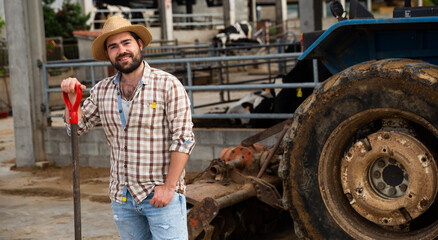 Confident young owner of dairy farm standing near tractor on background with cows in stall