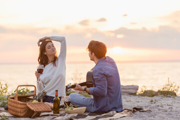 Selective focus of man playing acoustic guitar near girlfriend with wine on beach at evening