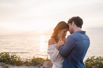 Young man touching brunette girlfriend on beach near sea at sunset