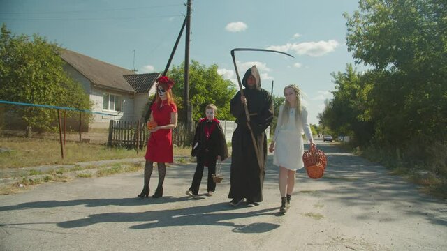 Diverse people and preadolescent boy with candy baskets in halloween make-ups and costumes, dressed as vampire, drowned woman, santa muerte and death trick-or-treating in village during halloween.