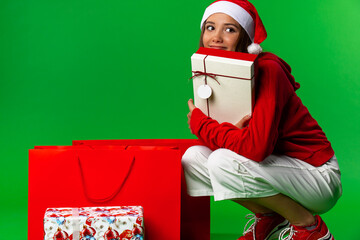 Smiling excited girl in red santa claus outfit holding christmas gift at her beard isolated on the green background