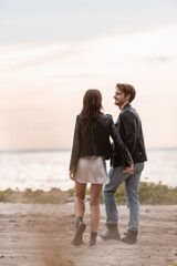 Selective focus of young couple in leather jackets holding hands while walking on beach at sunset