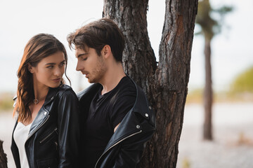 Brunette woman looking at boyfriend in leather jacket near tree in forest